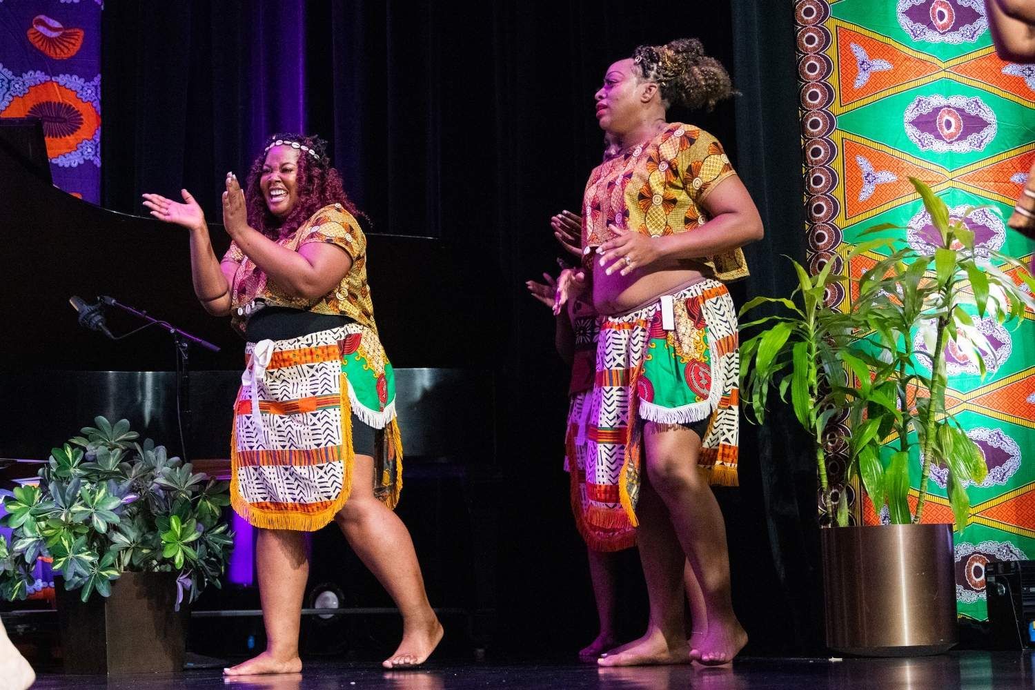 Two people in colorful African attire dance on stage. They clap and smile, with plants and patterned decor.