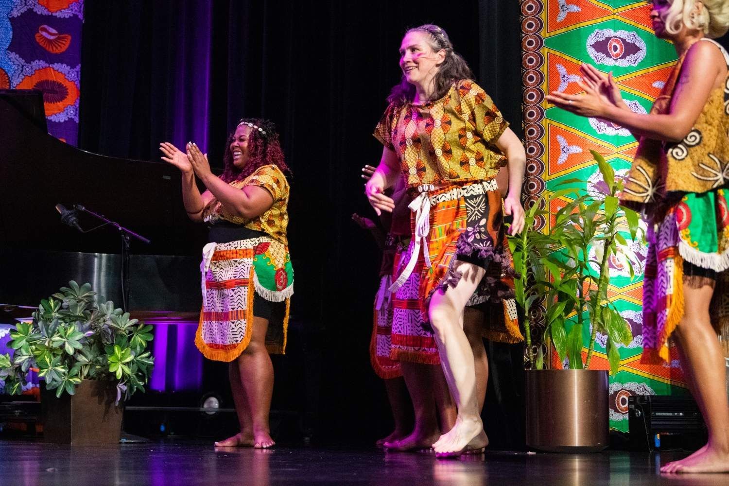 Three women in African-print outfits clap and dance onstage with a piano and colorful backdrop.