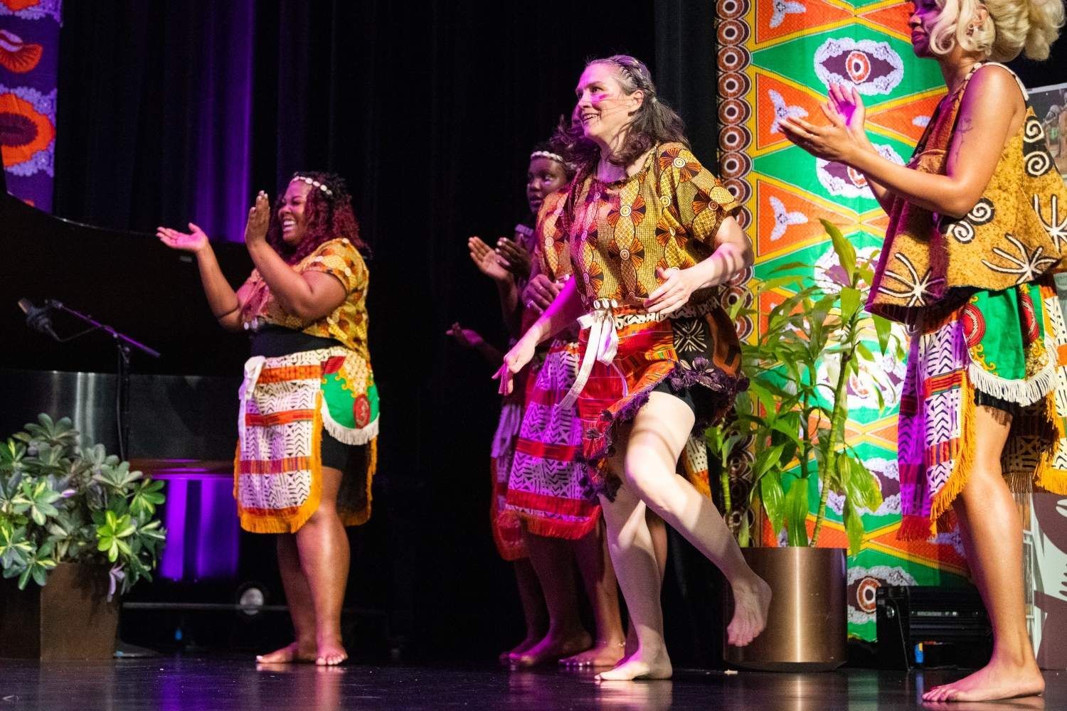 Group of women dancing on stage, wearing colorful African-style clothing. Barefoot, clapping, smiles, decorative backdrop.
