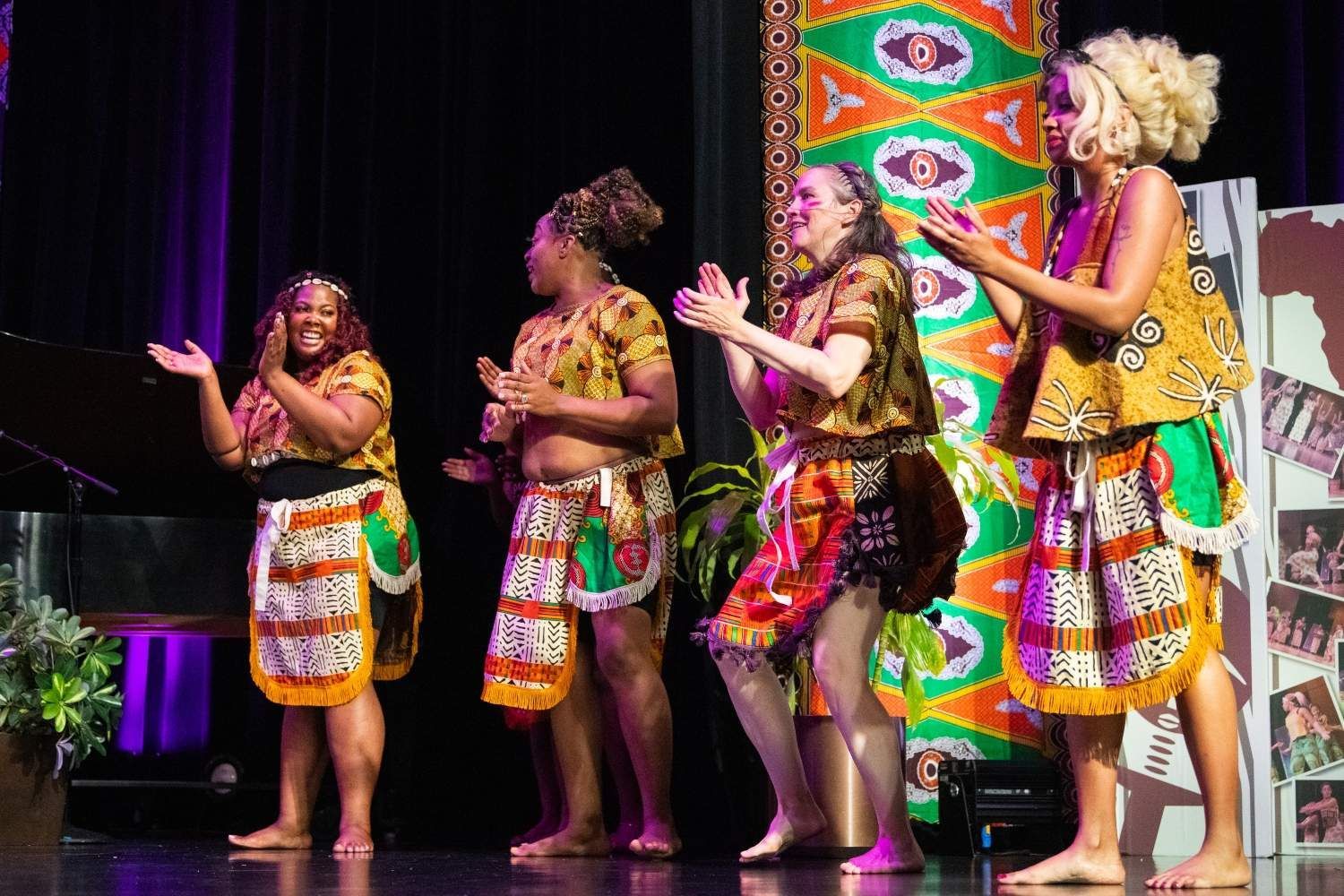 Four women on stage clapping, wearing African-inspired outfits in bright colors.