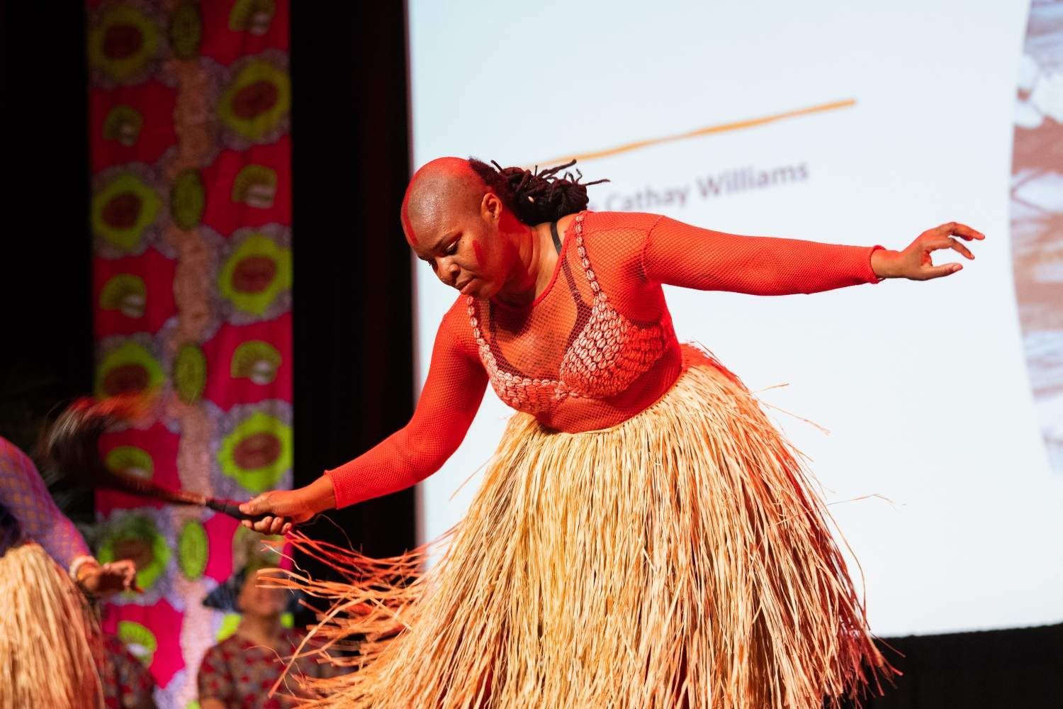 A woman dances on a stage, wearing a straw skirt and orange top, holding a stick.