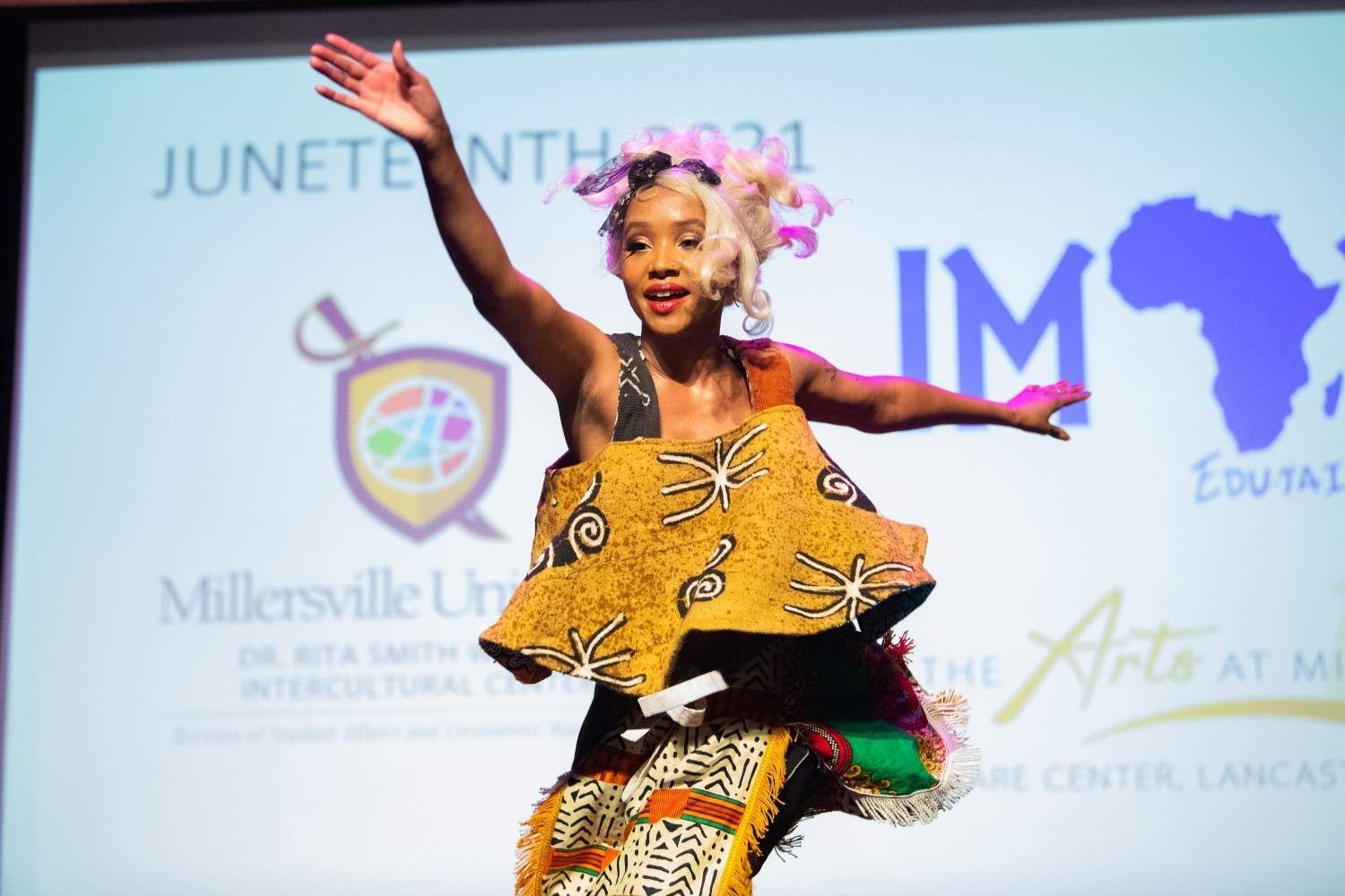 Woman on stage, arms up, wearing African-print clothing, in front of a Juneteenth 2021 backdrop.