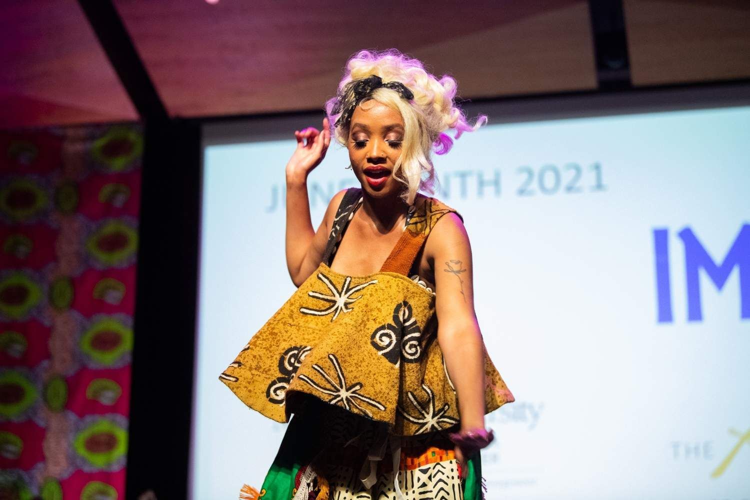 Woman in colorful African-print outfit on stage at Juneteenth event; curly blond hair, gesturing with hand.