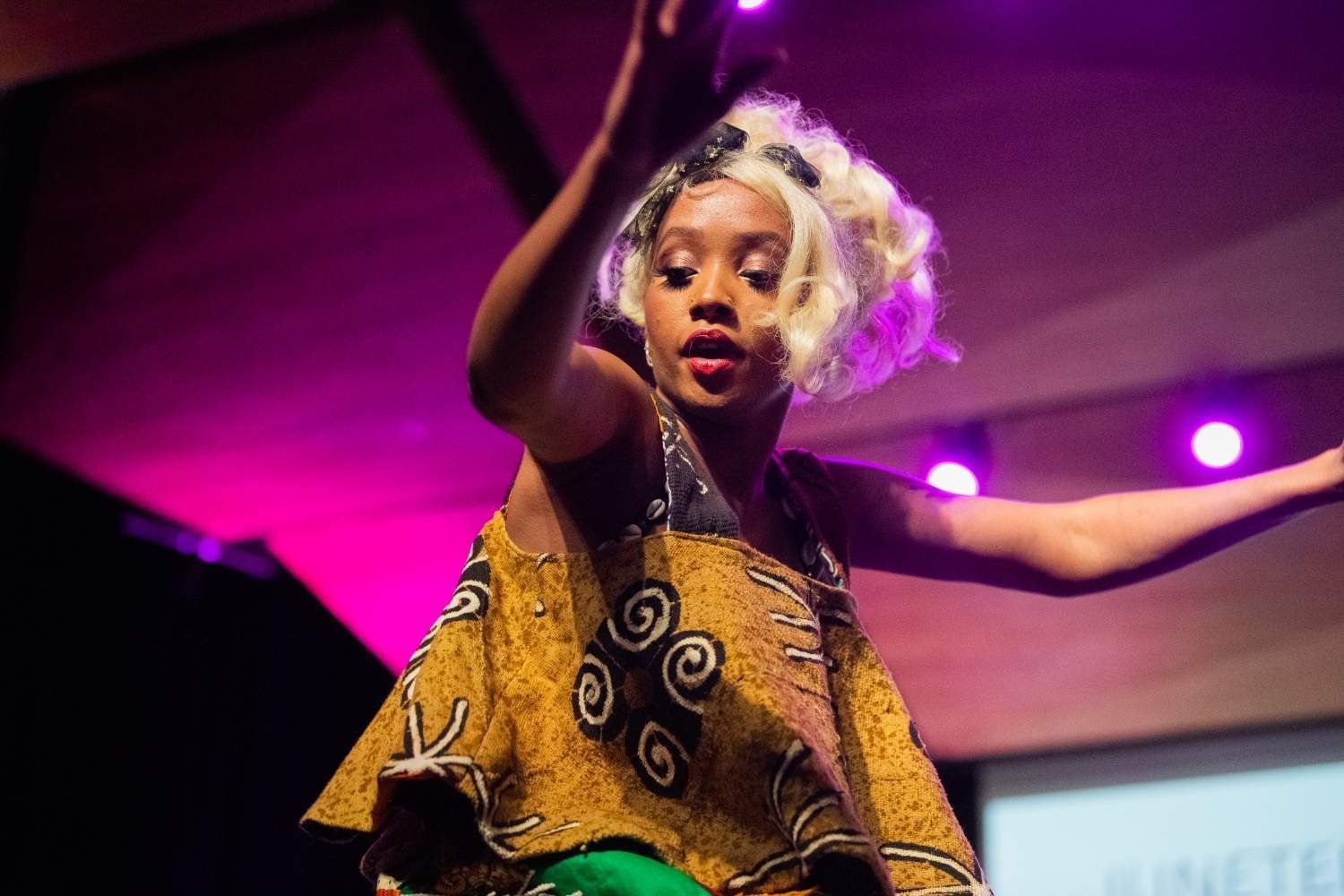 Woman in African-print top and blonde wig, dancing on a stage with pink and purple lighting.