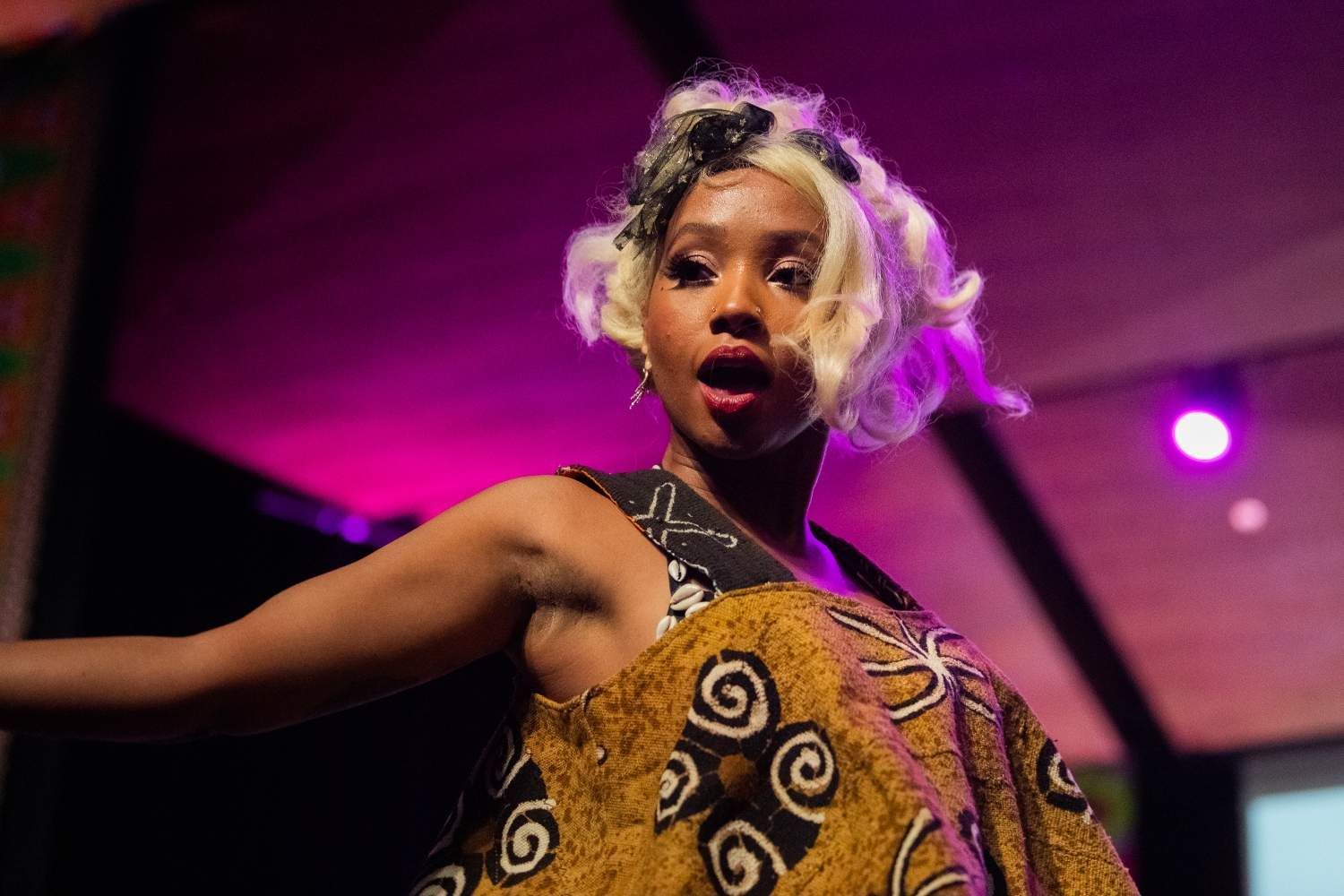 A Black person with black and white hair in performance wear with open mouth under stage lights.