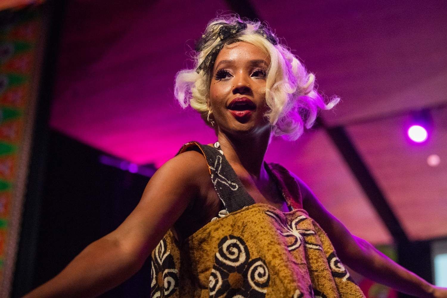 Woman in patterned dress, light hair, performing on stage with pink and purple lighting.