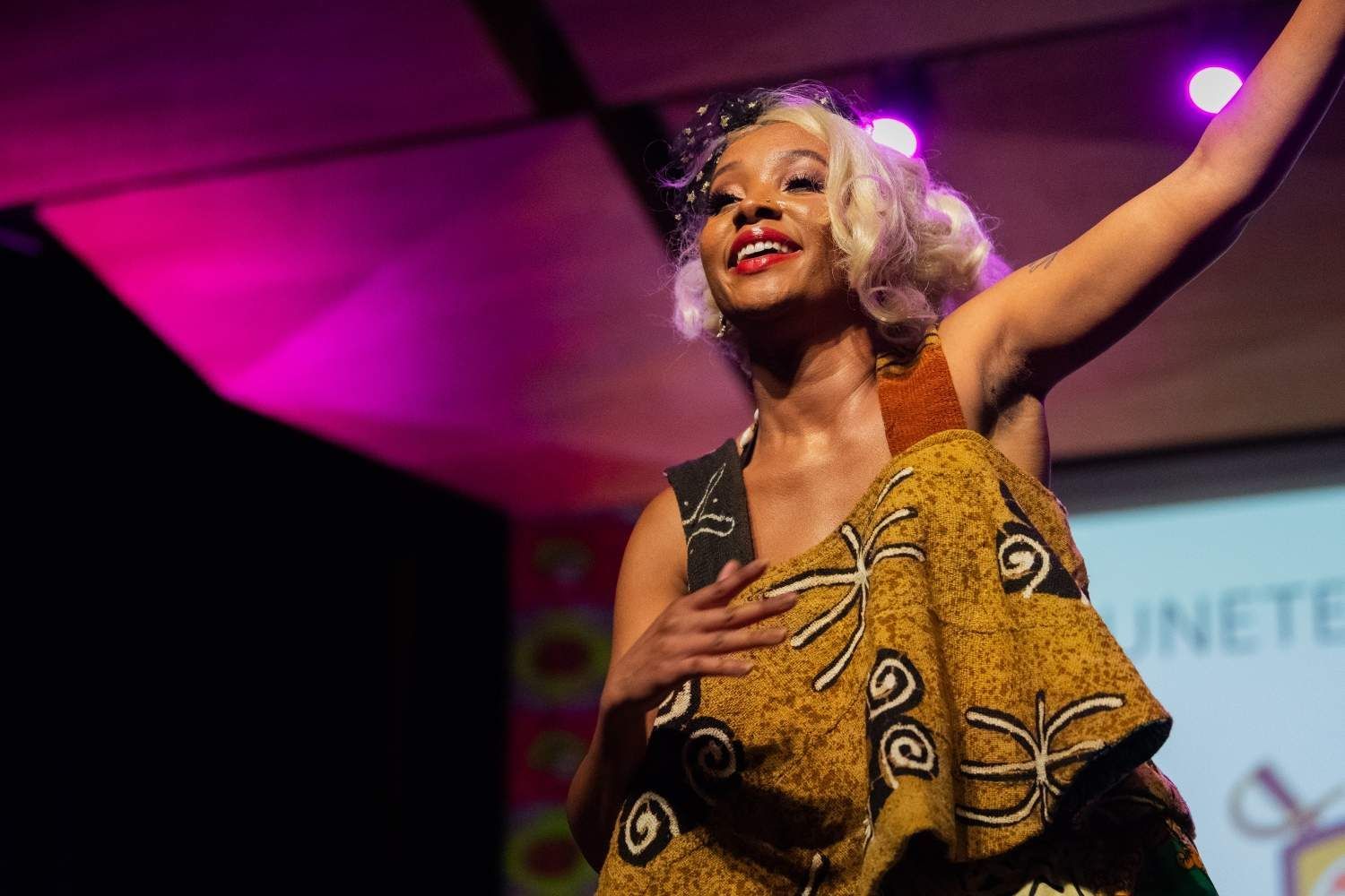 A Black woman with blonde hair, in a colorful dress, on stage, performing.