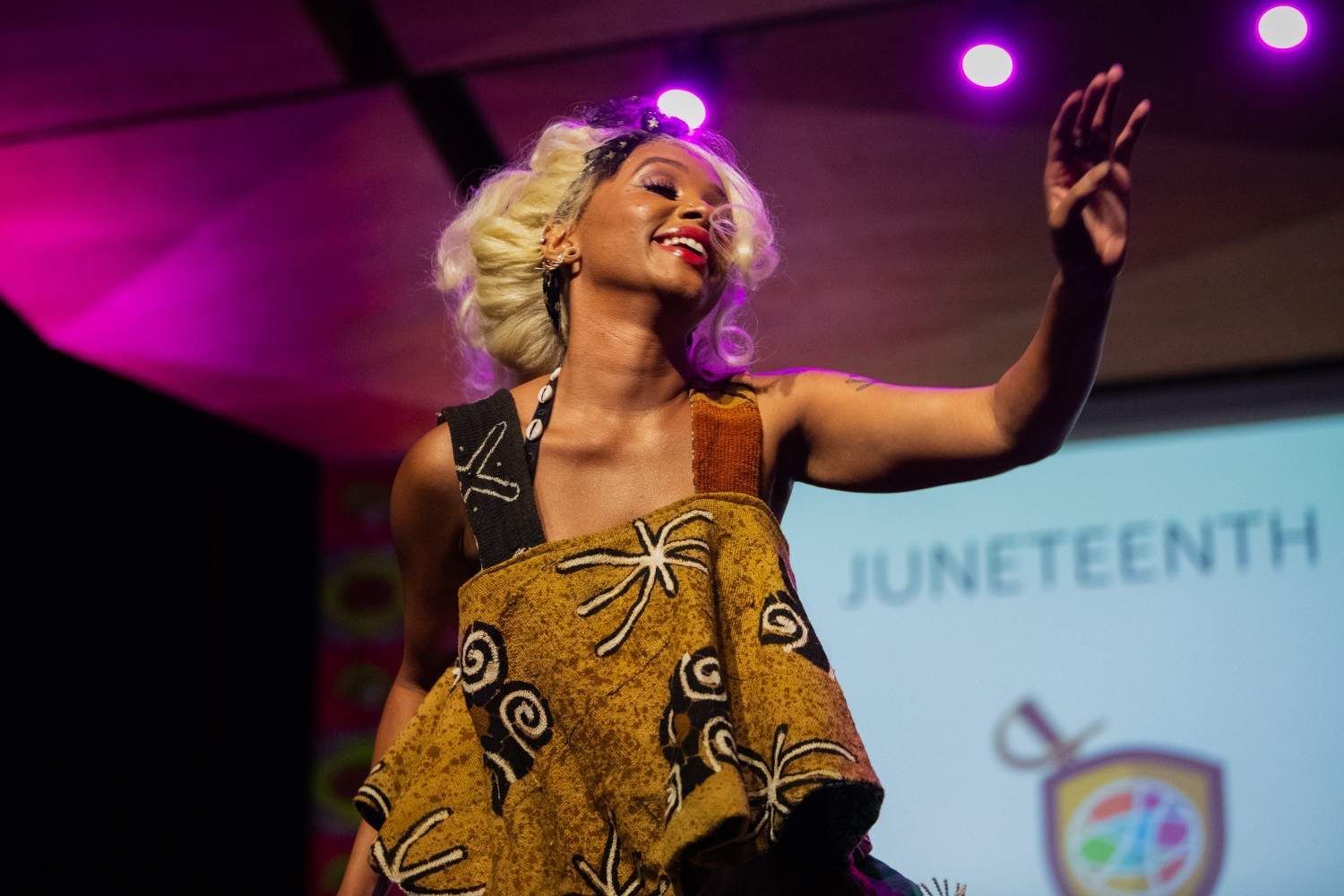 Woman in patterned dress waves on stage at Juneteenth event.