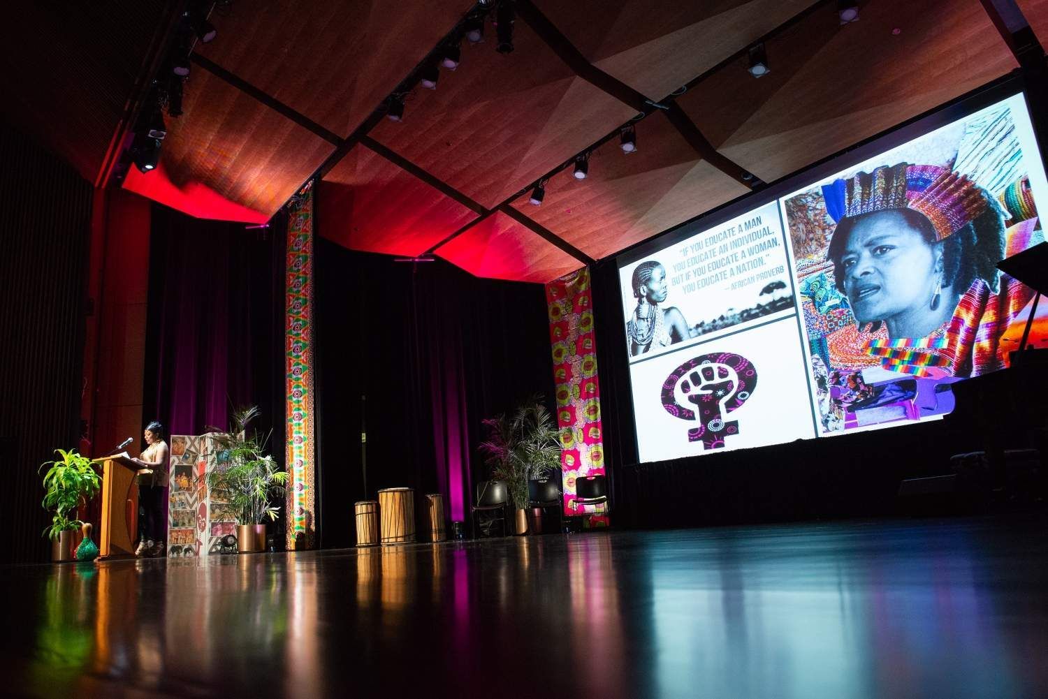 Woman at podium speaks on stage with slideshow of woman and feminist symbol.