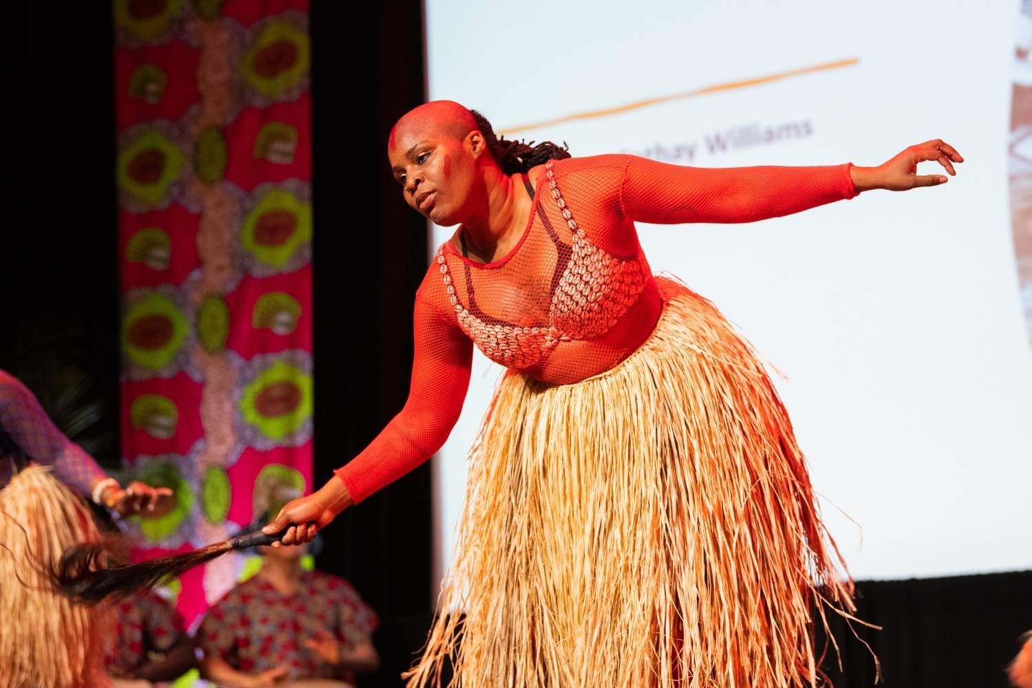 Woman in red top and straw skirt dancing onstage, holding a staff.