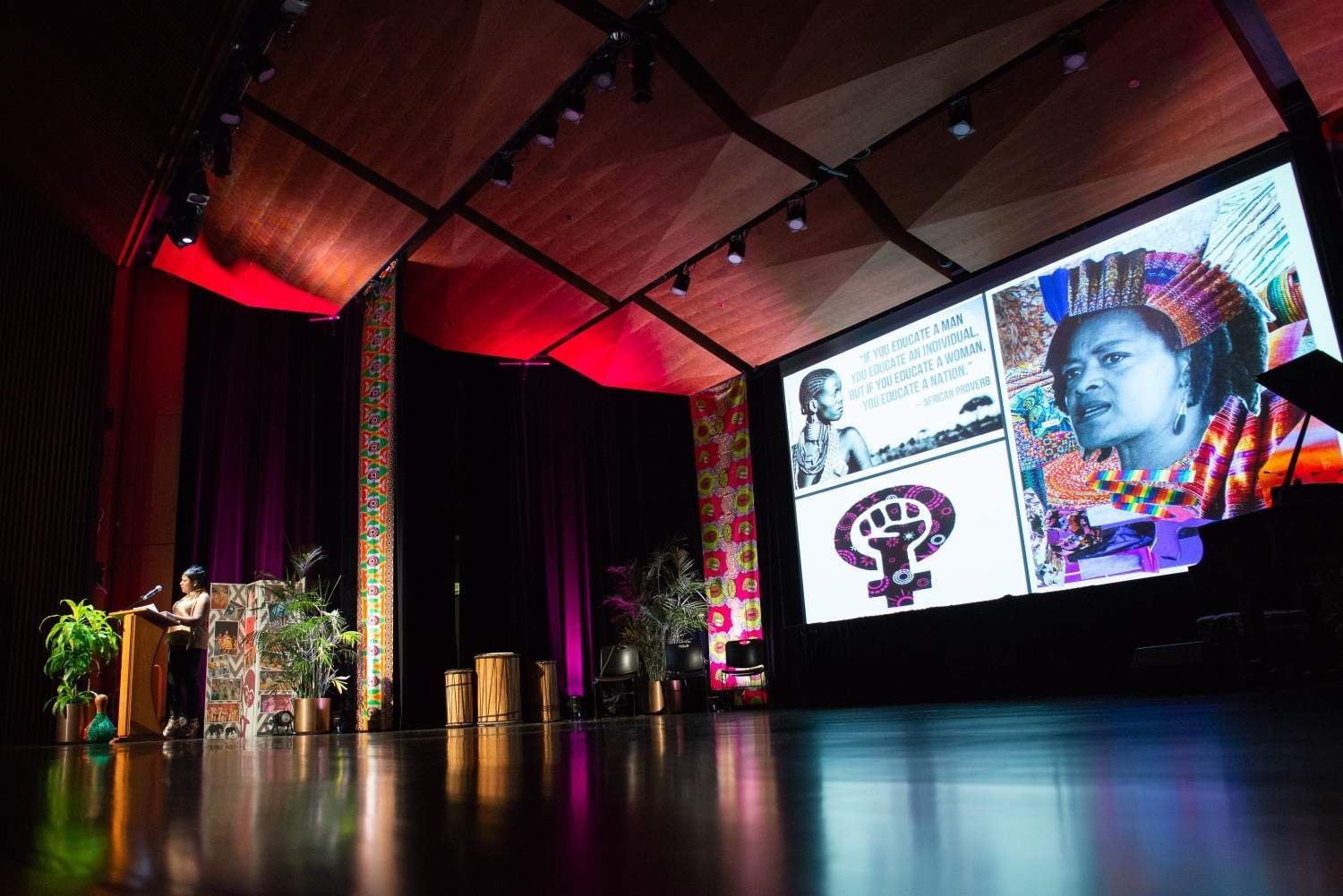 A woman speaks from a podium on a stage. A screen displays images and a feminist symbol.