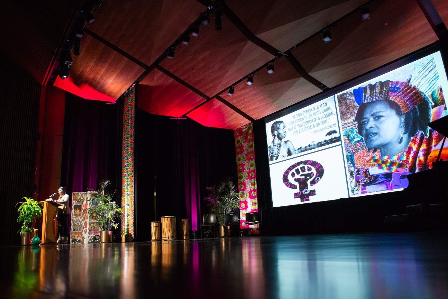 A speaker on a stage with a large screen displaying an image of a woman, in a decorated venue.