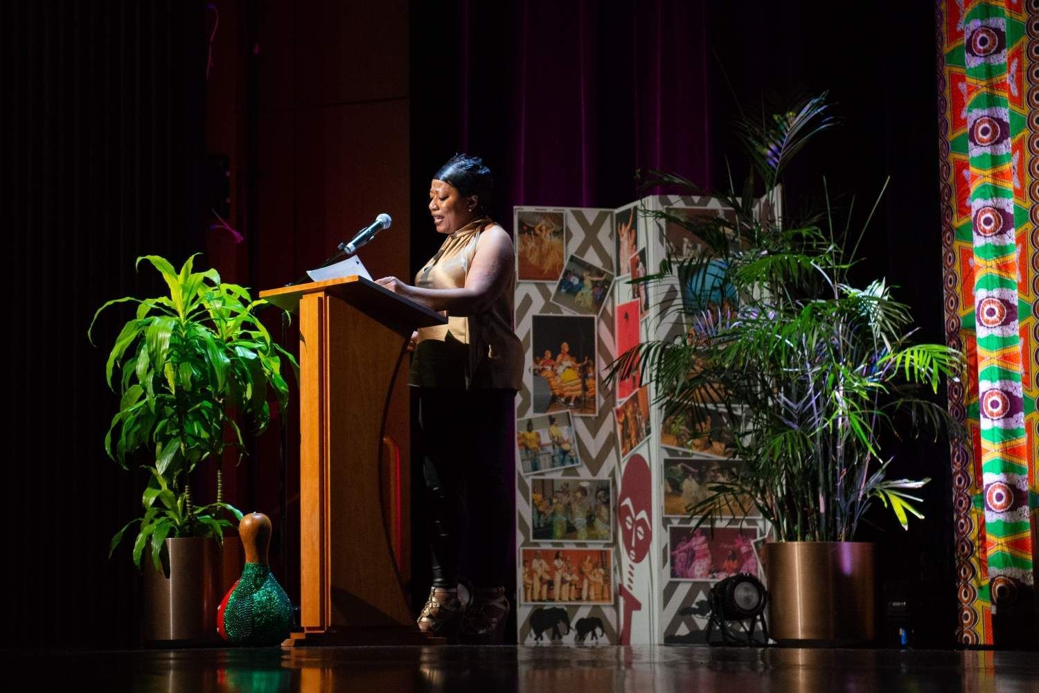 Woman speaking at a podium on a stage, next to plants and a decorative screen.