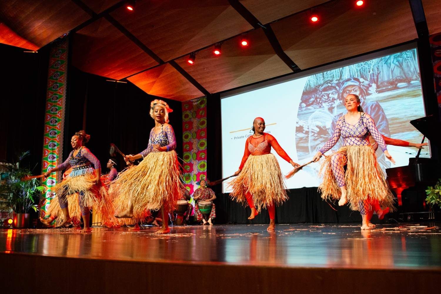 Dancers in colorful outfits perform on stage with a screen backdrop. They hold sticks and wear straw skirts.