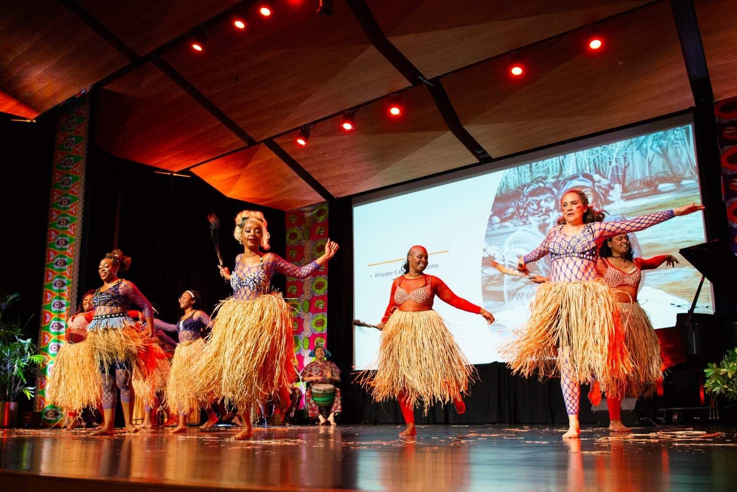 Dancers in colorful costumes perform on stage with a screen backdrop.