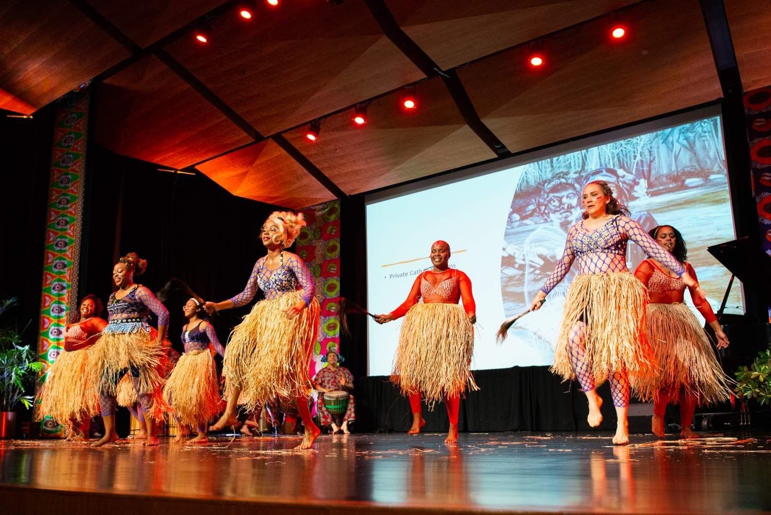 Dancers in colorful costumes perform on stage with a screen behind them.