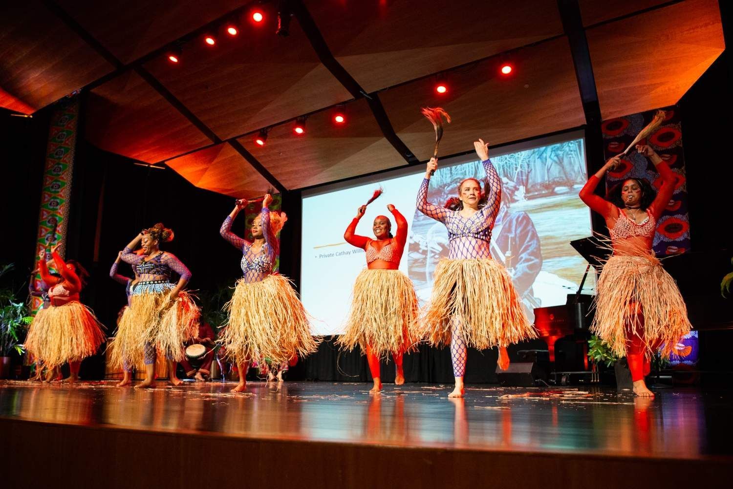 Dancers on a stage in colorful costumes with grass skirts, holding tools up. Stage lit with red.