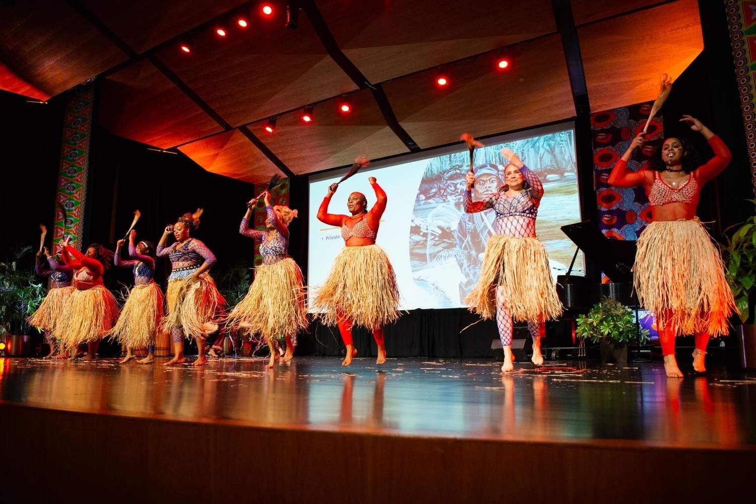 Dancers in straw skirts and red tops perform on stage, raising their arms.