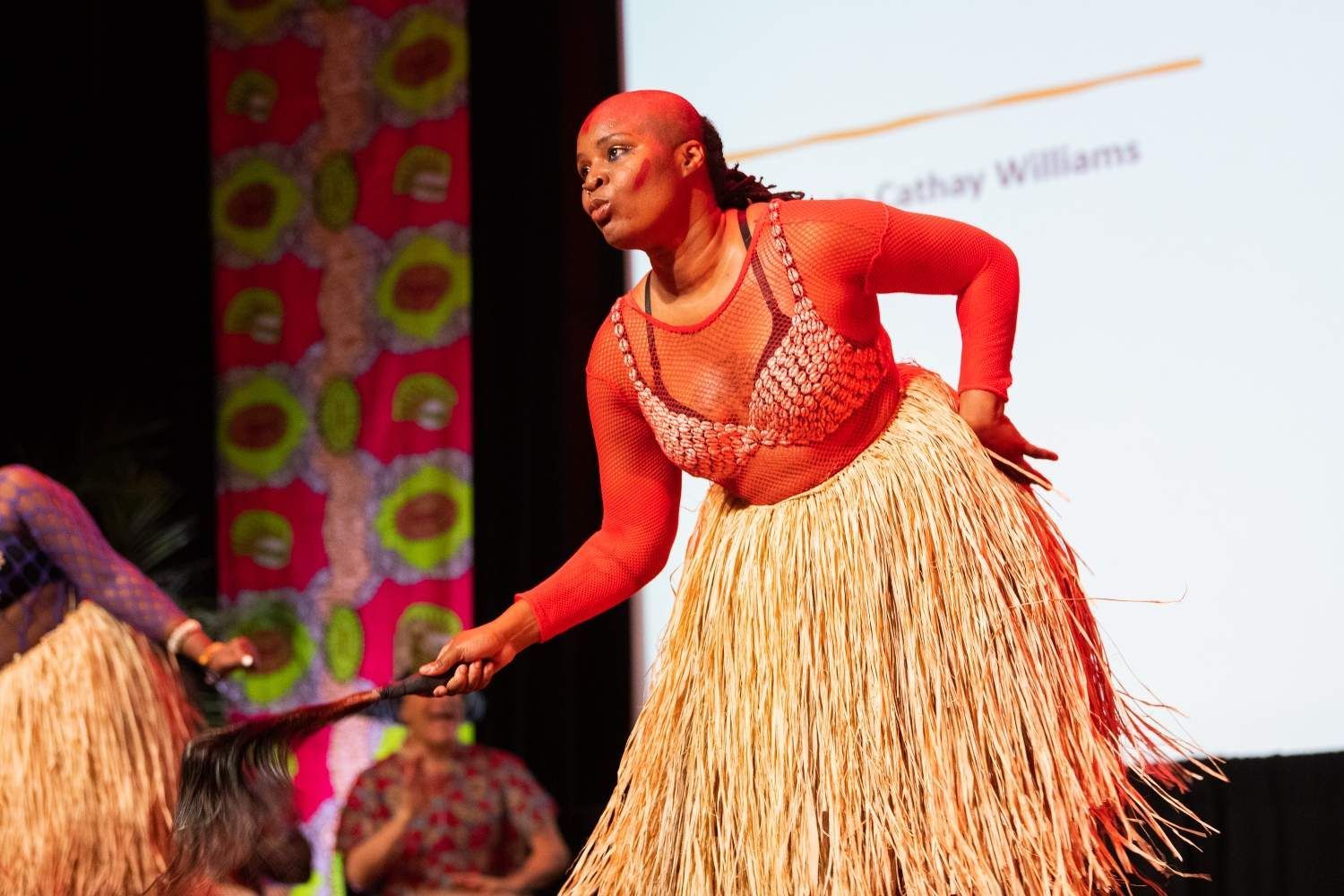 Woman in red top and straw skirt dances on stage, holding a staff.