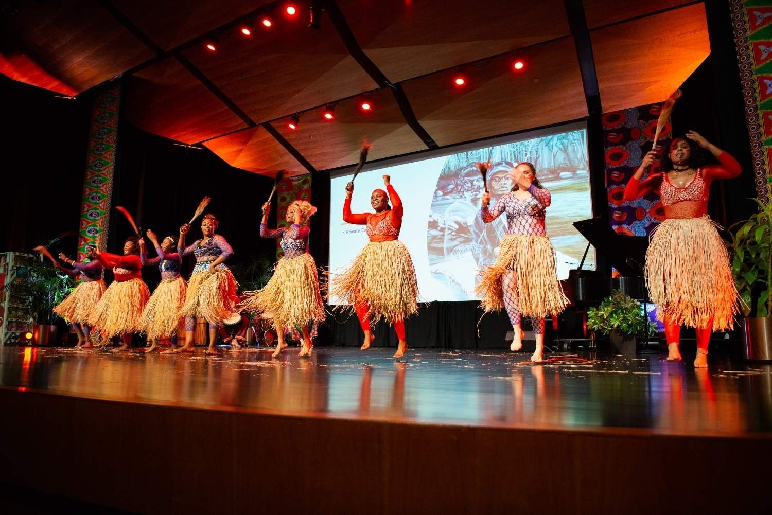 Dancers on stage in red tops and straw skirts, holding props. Lit backdrop.