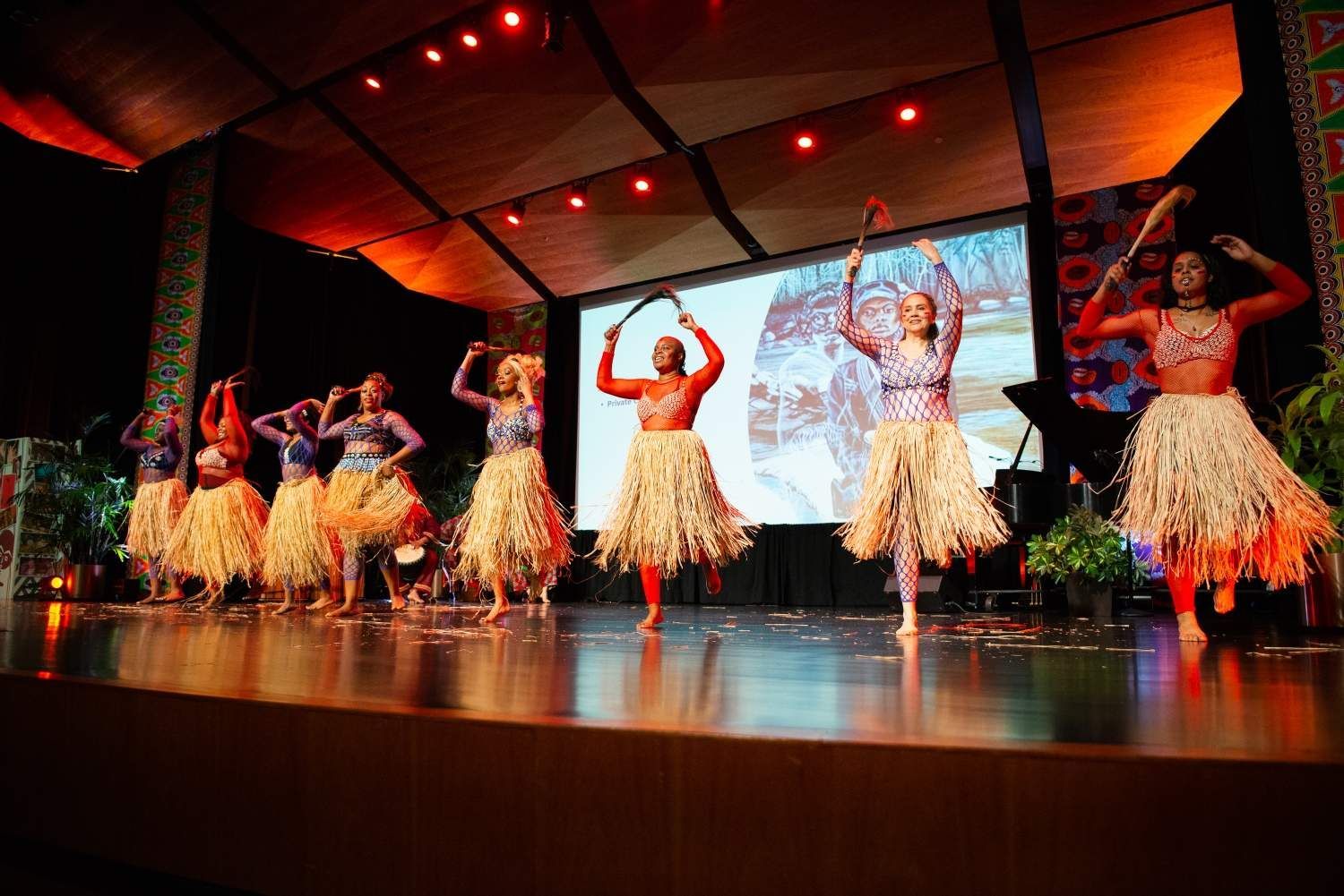 Dancers on a stage, wearing grass skirts, arms raised, performing in front of a screen. Lit with warm tones.