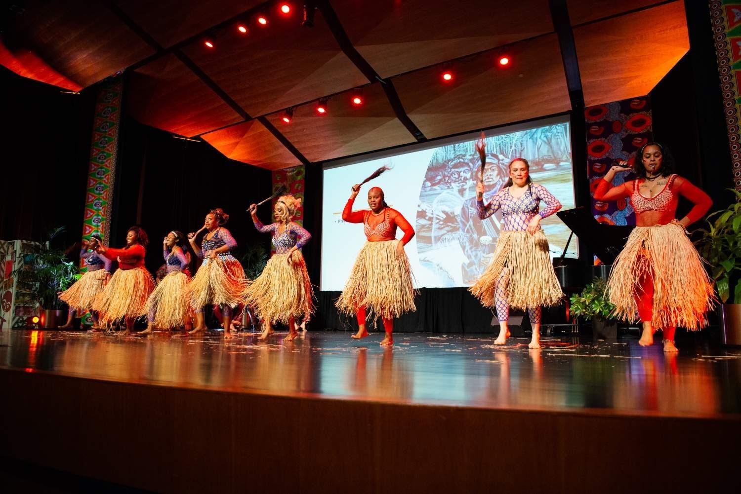 Group of dancers on stage in traditional attire, performing with props, lit by stage lights.
