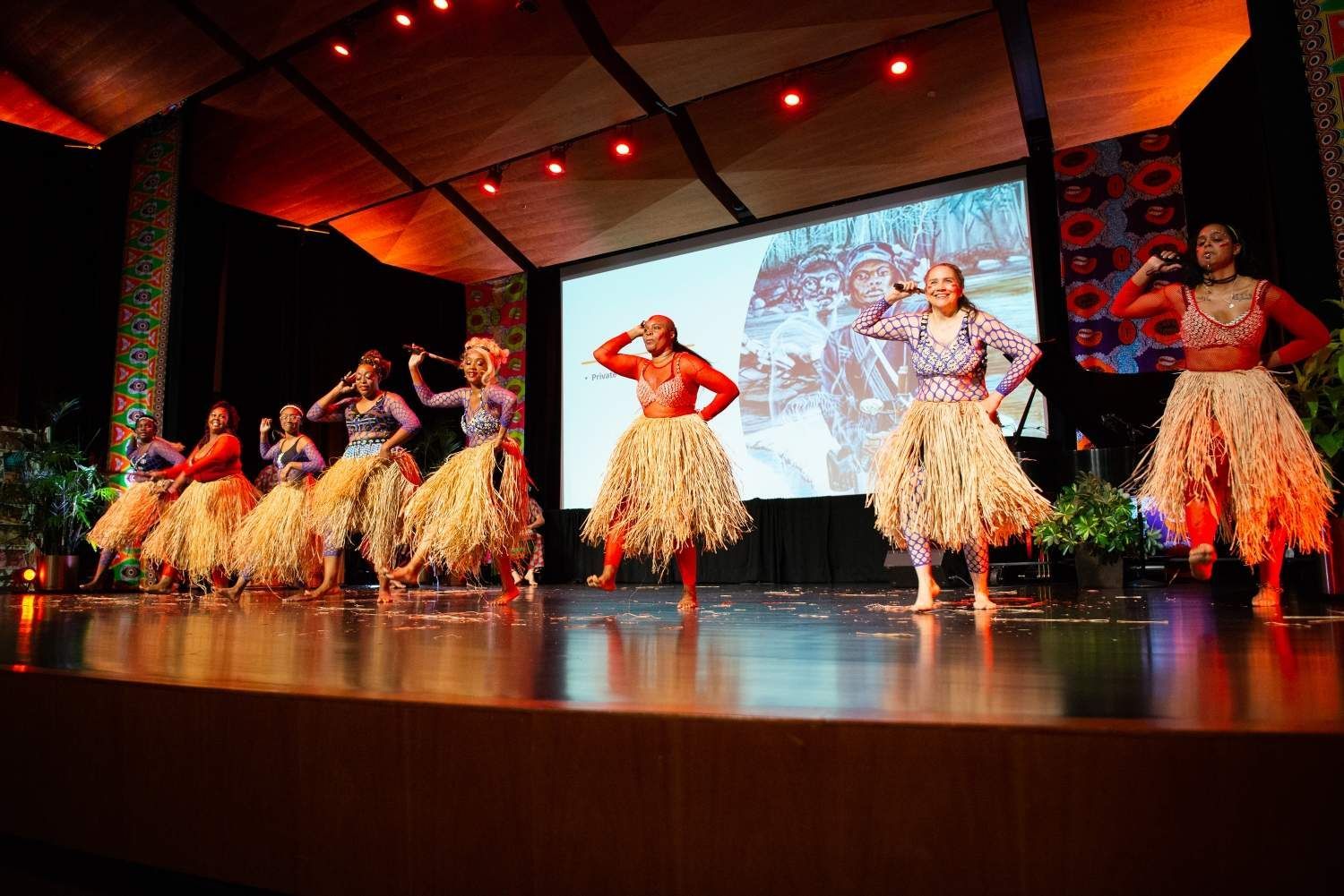 Dancers on stage in colorful tops and grass skirts, performing in front of a screen.