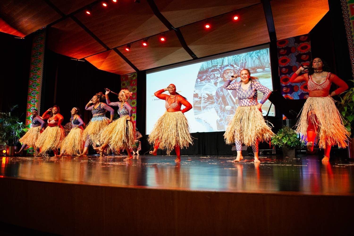 Dancers on a stage in colorful outfits and grass skirts perform, with a screen in the background.