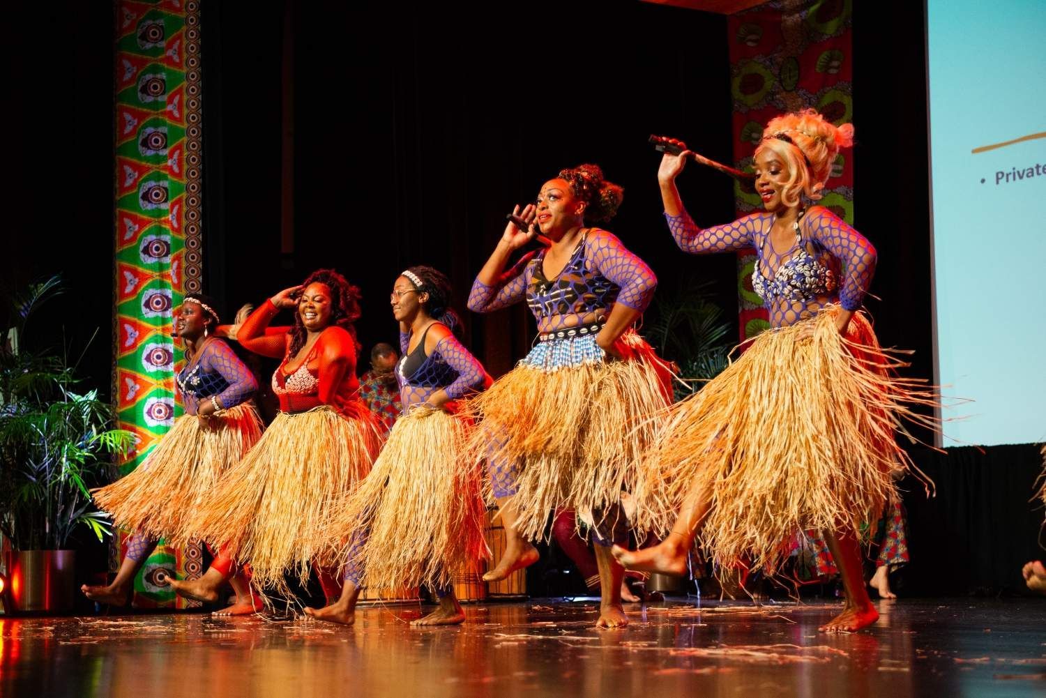 Five women in colorful outfits perform a dance on stage. They wear straw skirts and hold sticks.