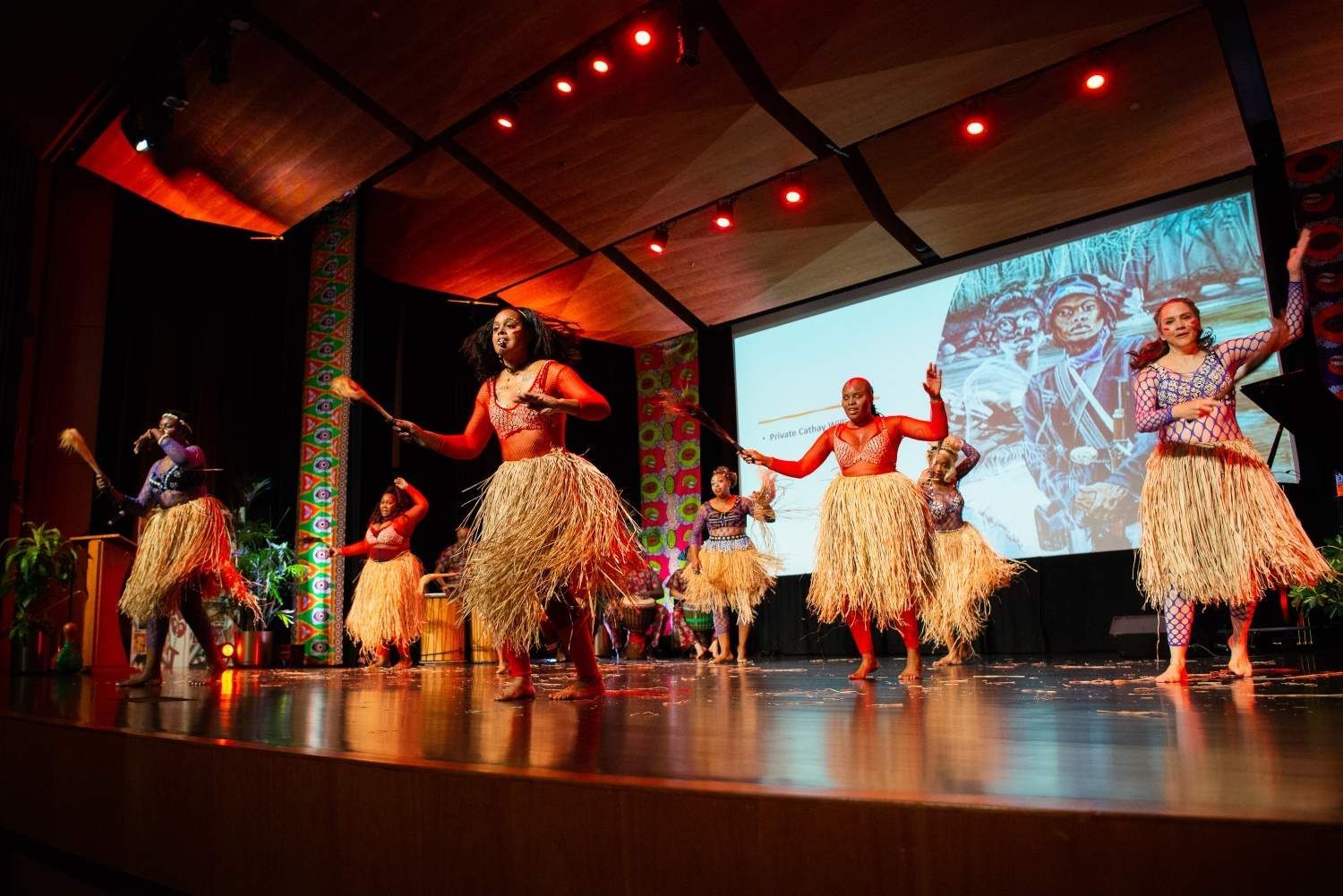 Dancers in grass skirts and painted bodies perform on stage with a screen in the background.