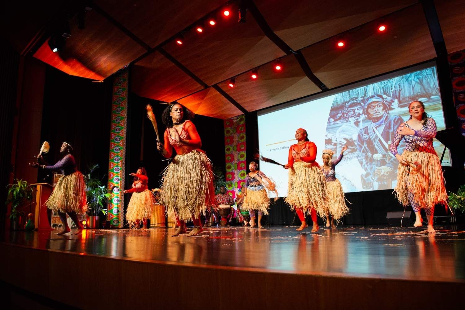 Dancers on stage in tribal clothing, performing with drums and props, projected background, stage lights.