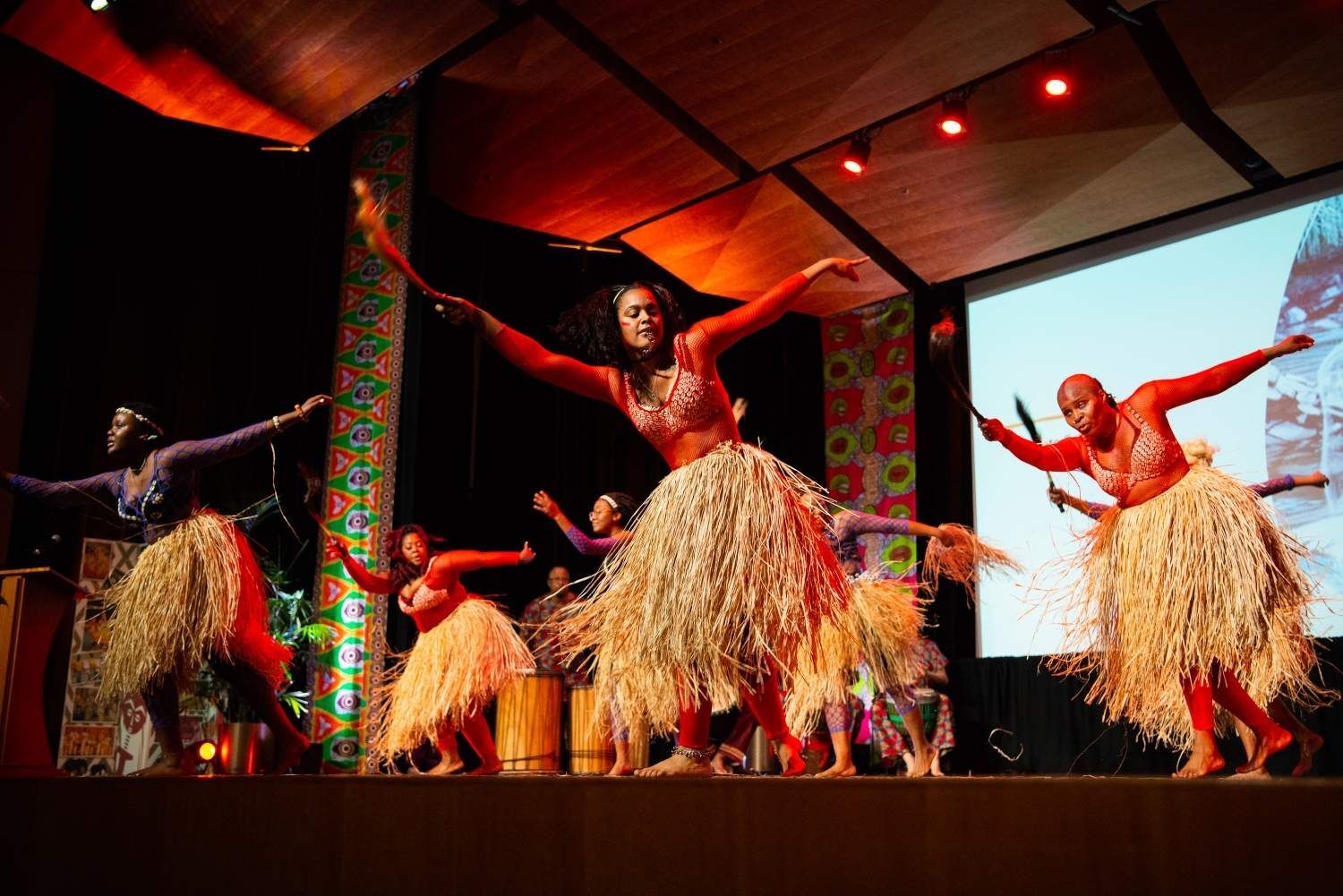 Dancers on stage in red body paint and grass skirts perform; stage has dark, colorful backdrop.
