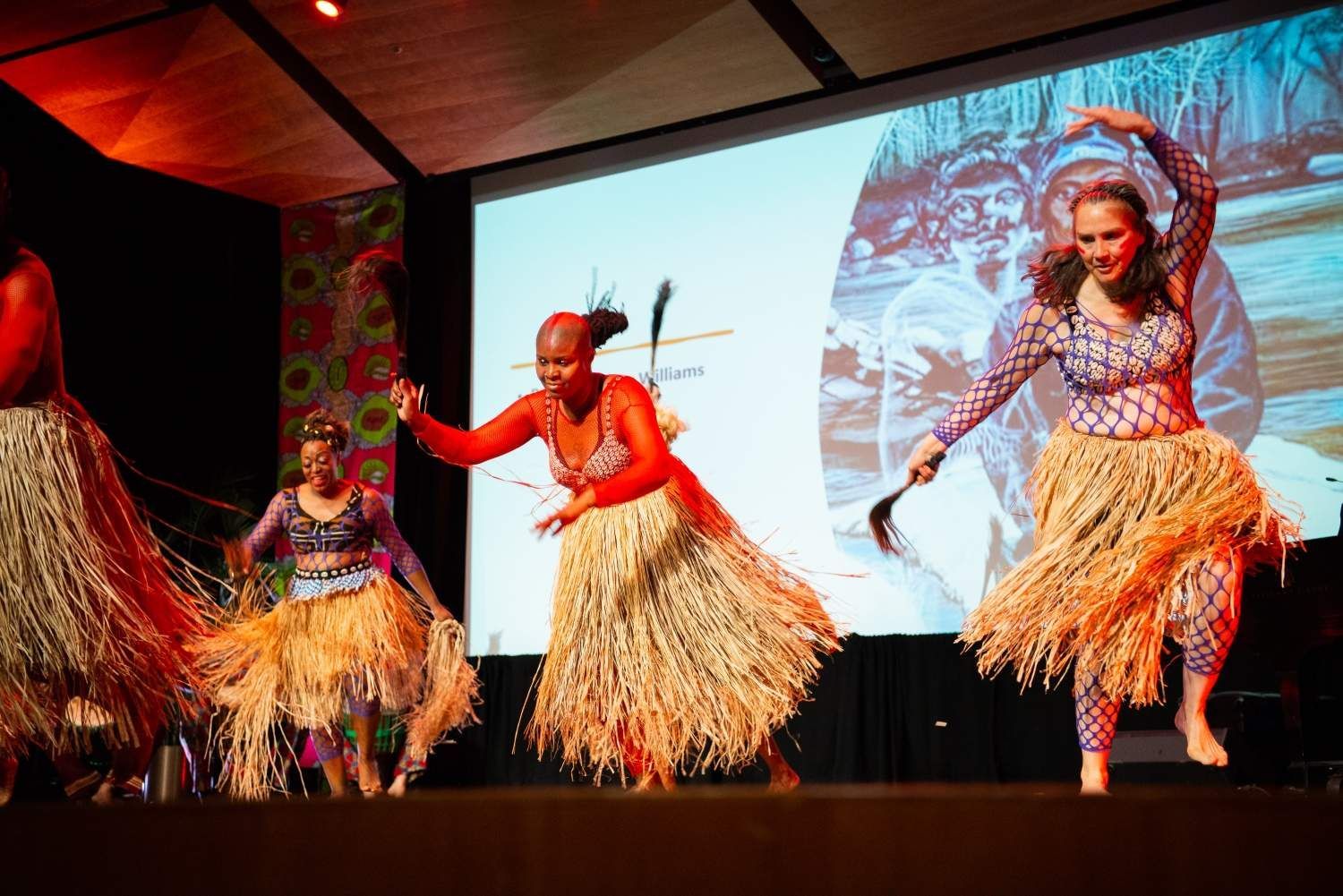 Dancers in colorful patterned tops and straw skirts perform on stage in front of a projected image.