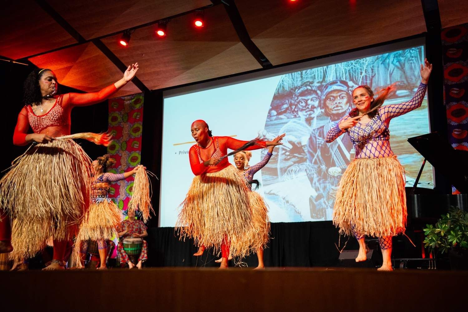 Dancers in grass skirts and colorful tops perform on stage, with a projected image in the background.
