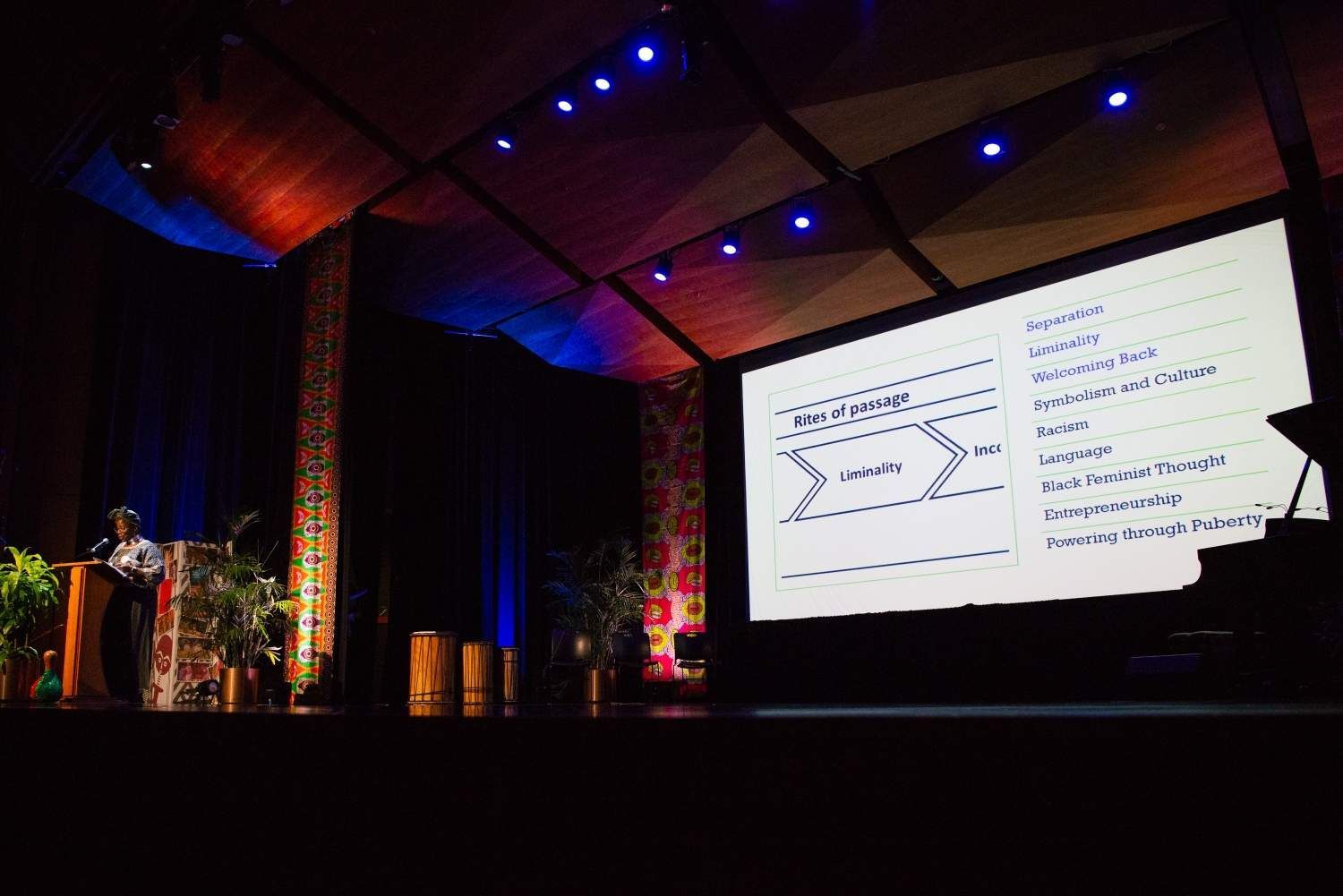 A presenter at a conference stands on stage, pointing to a slide with a diagram and text.