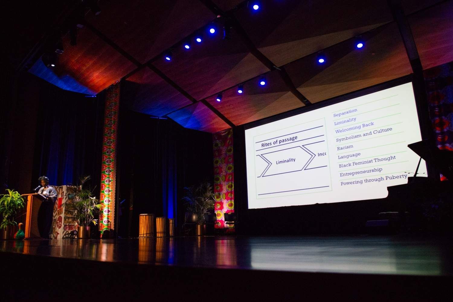 Speaker on stage with presentation screen, stage lit in blue and red.
