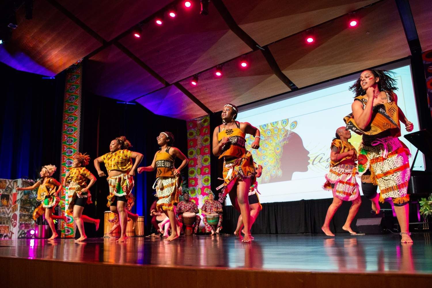 Group of dancers in colorful African-style outfits perform on stage; large screen in the background.