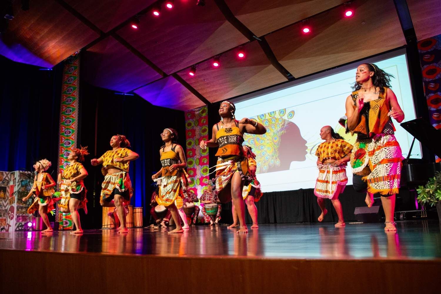 Dancers in colorful African attire perform on stage with projected backdrop.
