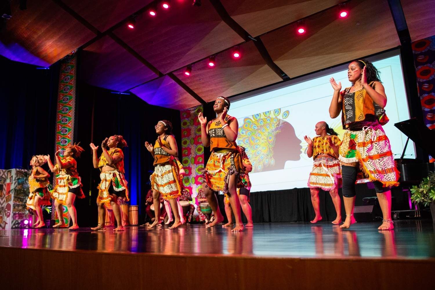 A group of people in colorful outfits dance on a stage, performing in front of a screen.