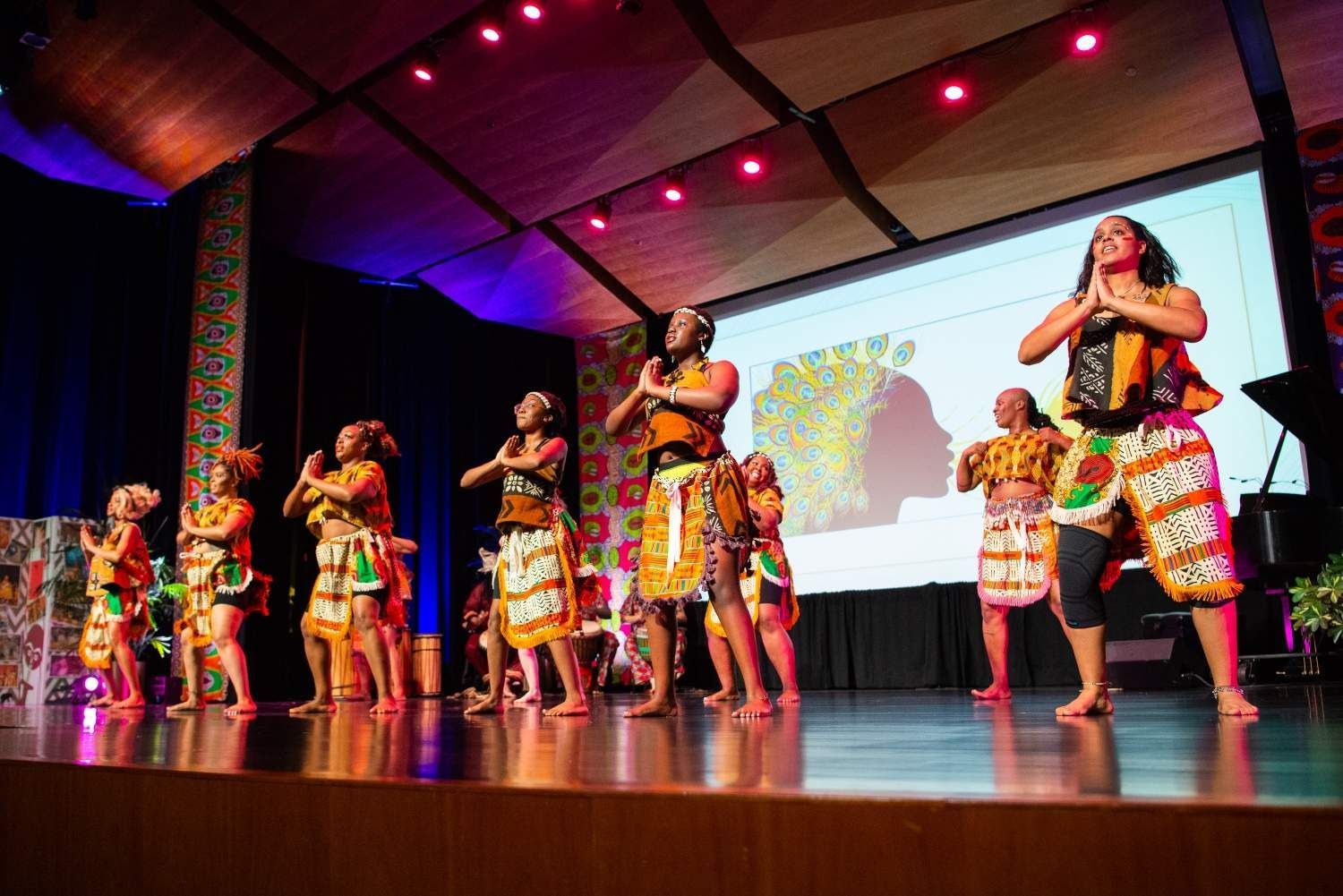 Dancers on stage performing, wearing colorful African-inspired outfits, with a projected image behind them.