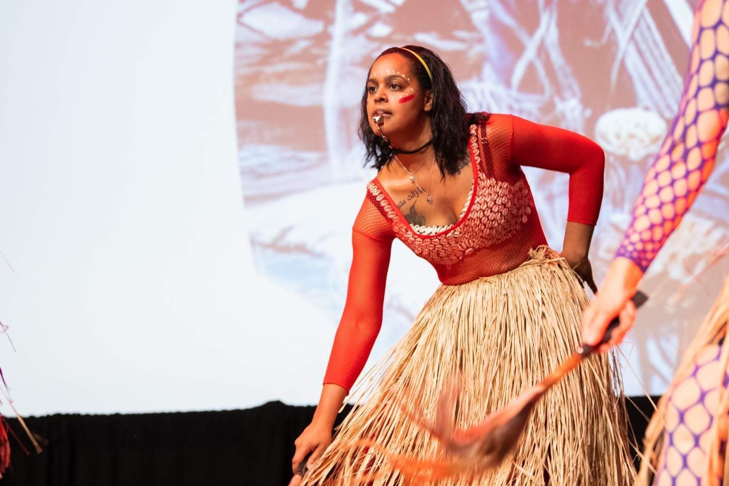 Woman in red top and straw skirt performs dance on stage.