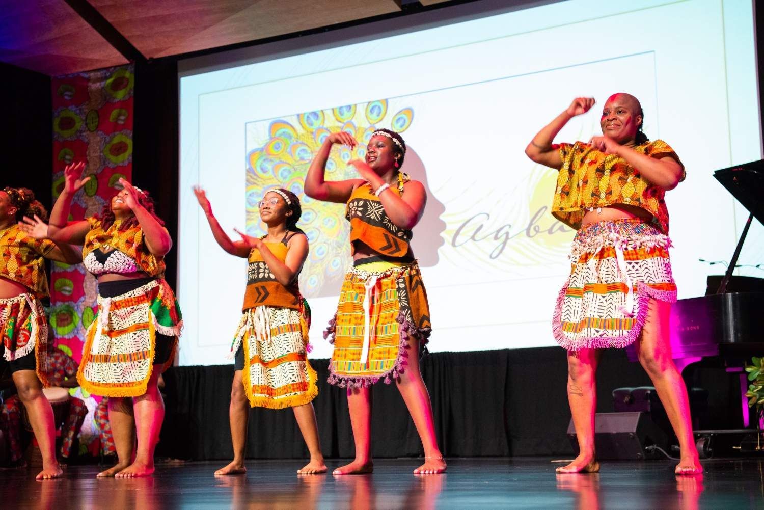 African dancers on stage, wearing patterned tops and skirts, performing with raised arms.