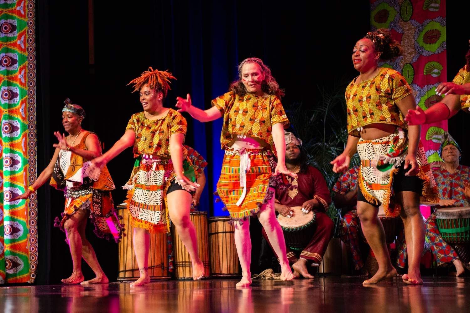 Dancers in yellow patterned tops and skirts perform on stage with drums, vibrant backdrop.