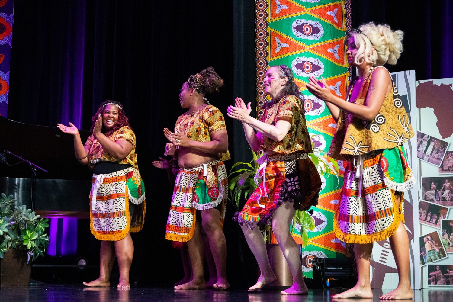 Four people in African-print outfits clap on stage. Backdrops include an African-print pillar, piano, and a map of Africa.