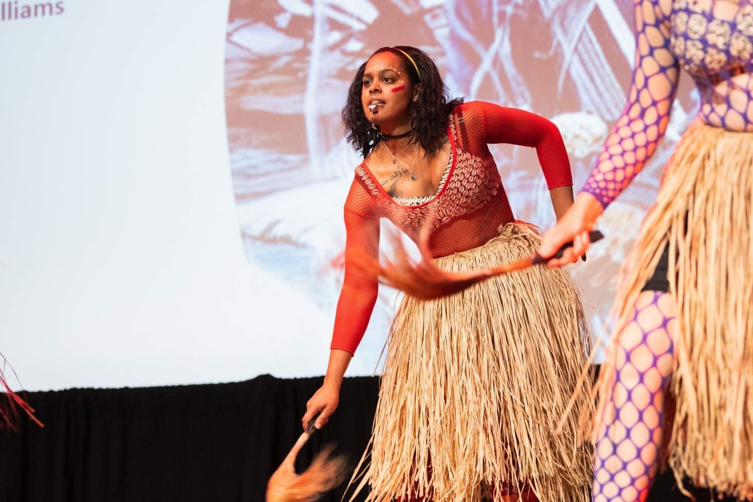 Woman in red and straw skirt dances on stage; others in background.
