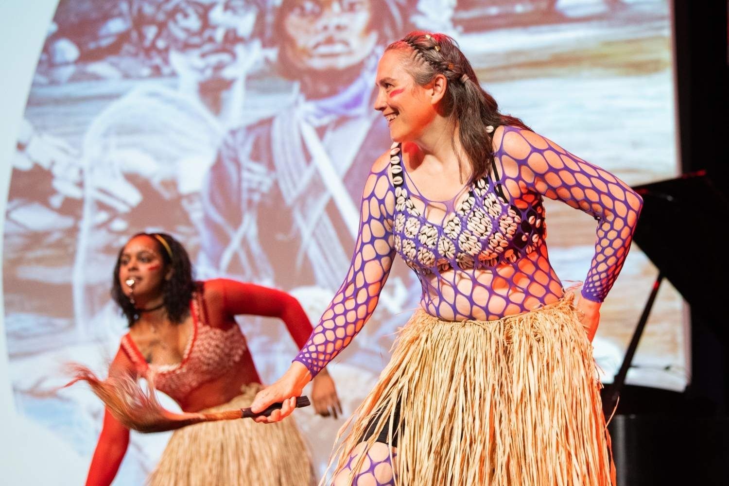 Two women dancing in grass skirts; background projection; smiling faces.