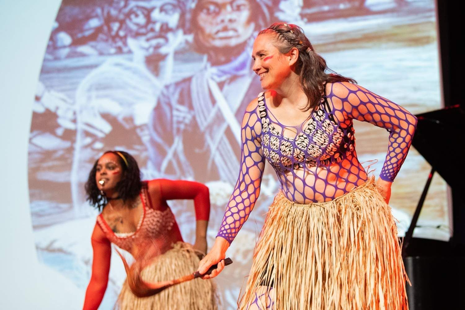 Two women dance onstage, wearing straw skirts and fishnet tops, with a projected image behind them.