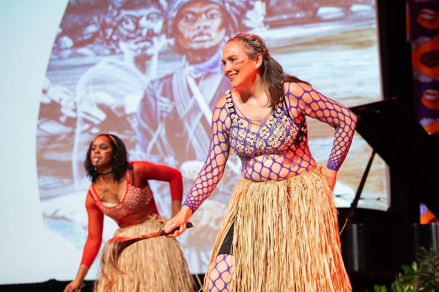 Two dancers in straw skirts and tops perform on stage. A backdrop shows a person in tribal garb.
