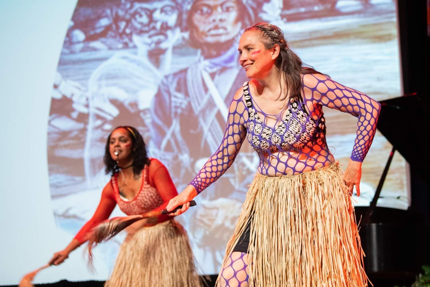 Two women in colorful costumes dance on stage with a projected background.