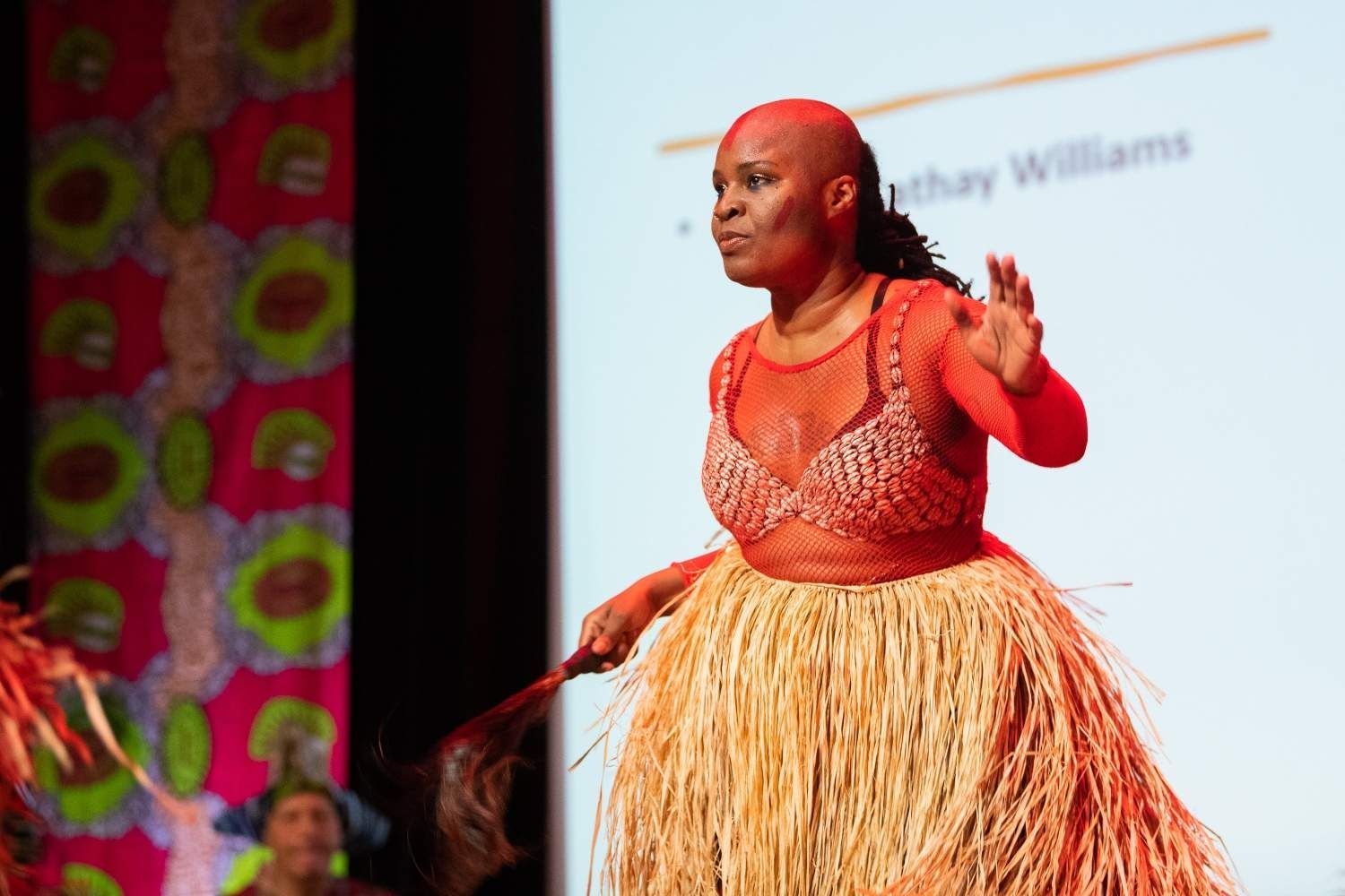 Woman in orange dress and grass skirt dances on a stage.
